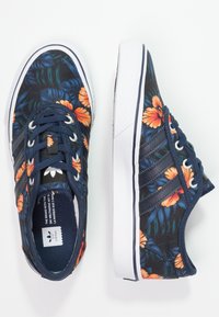 Marinblå kanvas-sneakers med blommönster, vita gummisulor, svarta sidestreck och ton-i-ton snörning. Accentuerade med orange blommor.