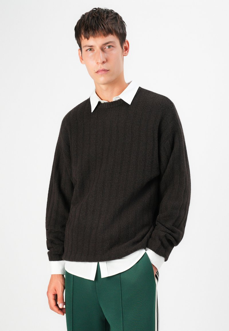 Pull en tricot côtelé marron foncé avec un col rond, porté par-dessus une chemise blanche à col. Coupe ample avec des poignets de manches retroussés.