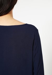 Marinblå topp med bred halsringning och långa ärmar, tillverkad av mjukt, strukturerat tyg, som visar baksidan av plagget.