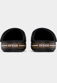 Ζευγάρι μαύρων Crocs χωρίς κορδόνια με καφέ και λευκές ρίγες και το λογότυπο των Crocs στον ιμάντα της φτέρνας, εμφανίζεται από πίσω σε λευκό φόντο.