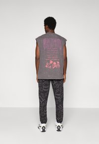 Mann med kort krøllete hår står med ryggen til iført ermeløs grå skjorte med rosa hodeskalle og "World Tour" tekst, mønstrede svarte bukser, hvite joggesko.