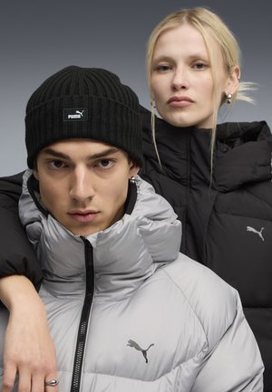 Two models wear puffer jackets; one in shiny silver, the other in matte black. Both feature high collars and prominent Puma logos.