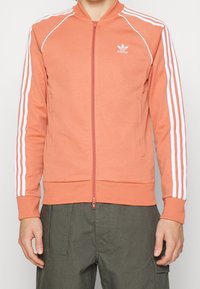 Orange zip-up jacka med vita vertikala ränder längs ärmarna, med ribbad krage och muddar, gjord av mjukt material.
