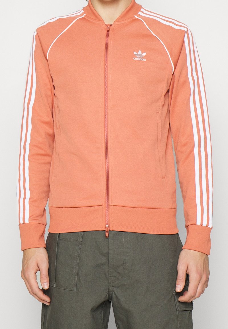 Orange zip-up jacka med vita vertikala ränder längs ärmarna, med ribbad krage och muddar, gjord av mjukt material.