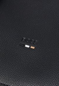Surface en cuir noir avec finition texturée, comportant un logo "BOSS" en relief et trois petites touches de couleur : noir, blanc et marron.