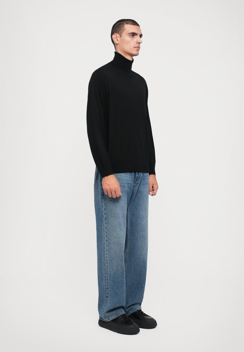 Schwarzer Rollkragenpullover aus weichem Stoff, kombiniert mit hellblauen, weit geschnittenen Jeans und schwarzen Schnürschuhen. Schlichte, moderne Optik.