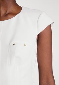 Weißes, figurnahes Kleid mit kurzen Ärmeln, das eine glatte Textur aufweist, goldene Hardware-Akzente an den Taschen und strukturierte Nähte zur Konturierung.