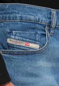 Haalistuneet siniset farkut, joissa näkyy takatasku, jossa on punavalkoinen Diesel Industry Denim Division -etiketti ja oranssit ompeleet.