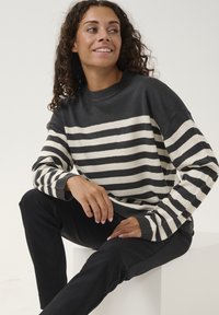 Grau-cream gestreifter Pullover mit geribbeltem Ausschnitt und Bündchen. Das Model trägt ihn mit schwarzen Hosen und sitzt auf einem weißen Podest.