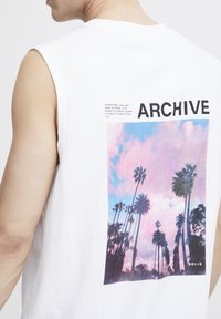 Fehér ujjatlan póló, hátul színes pálmafa grafikával, felette a "ARCHIVE" szó vastag fekete betűkkel.