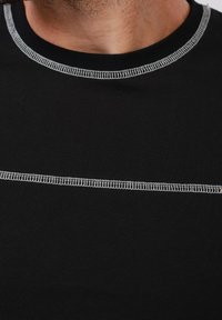 Κοντινό πλάνο του λαιμού και του άνω μέρους του στήθους ενός ατόμου που φορά μαύρο πουκάμισο με λευκή ραφή overlock στον γιακά και κατά μήκος του στήθους.