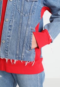 Džínová bunda s třepícím se lemem, s červenými pletenými detaily na manžetách a bocích. Design zahrnuje dvě náprsní kapsy a modrý prací efekt.
