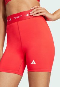 Czerwone sportowe szorty z wysokim stanem o gładkiej teksturze, z białym logo na pasku i dolnej części nogawki.