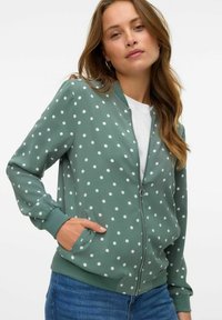 Veste bomber verte à pois blancs, fermeture éclair sur le devant, poignets et ourlet côtelés, poches latérales, portée par-dessus un T-shirt blanc.
