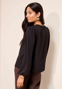 Hosszú sötét hajú nő fekete hosszú ujjú blúzban, arany fülbevalóval, nyaklánccal és karkötővel áll egy sima bézs fal előtt.