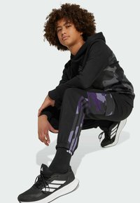 Black hoodie with textured camouflage, paired with black sweatpants featuring purple camouflage accents and three white stripes, black sneakers.