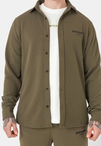 Camicia a maniche lunghe verde oliva con chiusura a bottoni, tessuto testurizzato e stampa del logo. Abbinata a una maglietta bianca sottostante.
