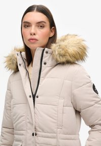 Polstret lyse beige jakke med en hætte foret med pels, lynlåslukning, trykknapper og en patch på ærmet. Har en quiltet struktur.