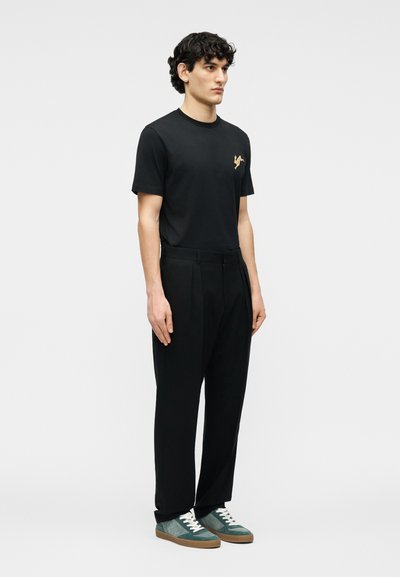 Άνδρας μοντέλο στέκεται φορώντας μαύρο μπλουζάκι με χρυσό λογότυπο, μαύρο πλισέ παντελόνι και πράσινα σουέντ αθλητικά παπούτσια με λευκά κορδόνια.