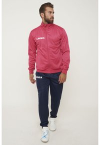 Rosa idrottsjacka med hög krage och vit logga, ihop med marinblå joggingsbyxor. Vita sneakers med blå detaljer fulländar looken.