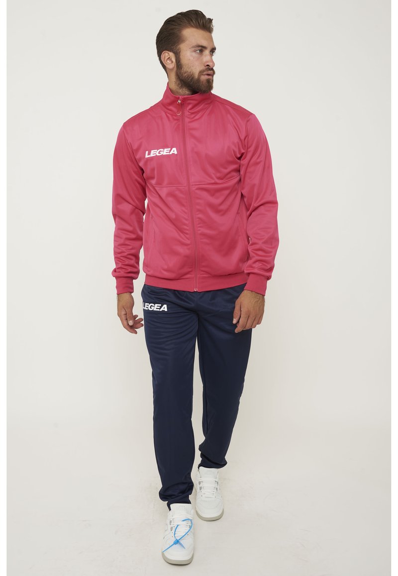 Rosa idrottsjacka med hög krage och vit logga, ihop med marinblå joggingsbyxor. Vita sneakers med blå detaljer fulländar looken.