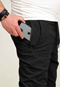 Black joggers with a textured finish, featuring a zippered side pocket holding a gray smartphone. Simple design with drawstring waist.