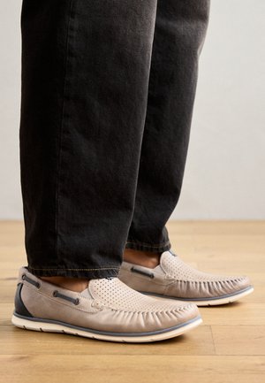 Beżowe buty slip-on wykonane z miękkiej skóry, z dziurkami, dekoracyjnym przeszyciem oraz kontrastującym granatowym akcentem na pięcie.