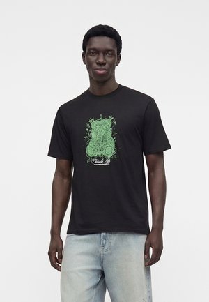 Homme portant un t-shirt noir avec un motif ours en peluche vert et un jean bleu clair, debout devant un fond blanc uni.