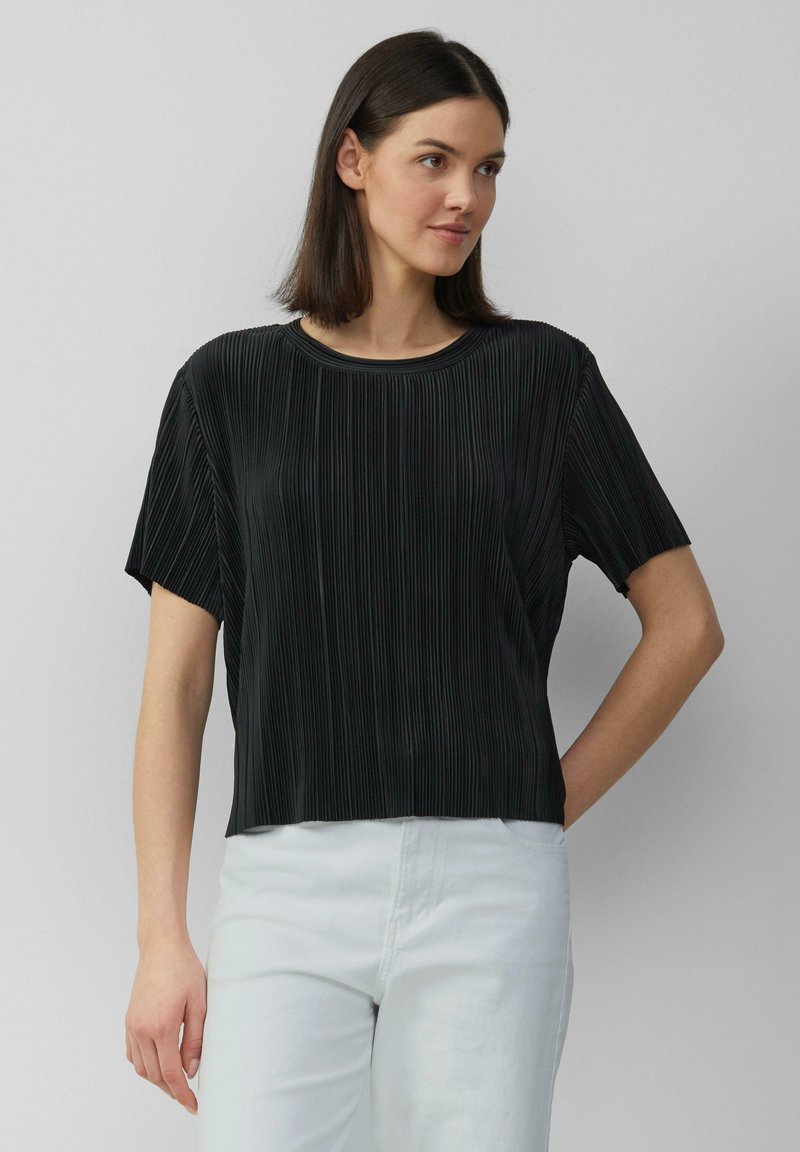 Czarna plisowana bluzka z krótkim rękawem i okrągłym dekoltem, wykonana z miękkiego materiału, zestawiona z białymi spodniami, na neutralnym tle.