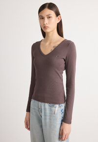 Bluză cu mânecă lungă, de culoare marron ciocolată, cu decolteu în V și un logo mic brodat. Asortată cu blugi relaxați de culoare albastru deschis.