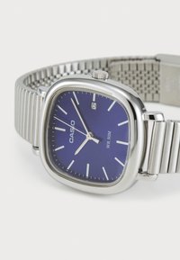 Montre en acier inoxydable argenté avec un cadran bleu carré, des index horaires blancs et un affichage de la date à 3 heures. Elle dispose d'un bracelet en maille.