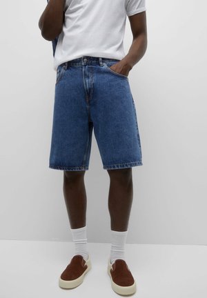 Un homme porte un short en denim bleu mi-long, des chaussettes blanches, des chaussures marron à enfiler, et un t-shirt blanc rentré dans le pantalon, une main dans la poche.
