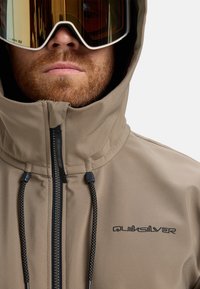 Bežna skijaška jakna s visokim ovratnikom, prednjim zatvaračem, crnim konopcima i Quiksilver logom. Model nosi reflektivne skijaške naočale.