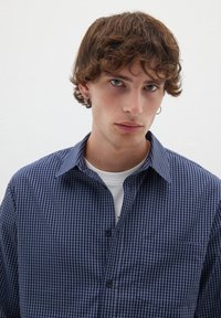 Camicia a quadri blu e bianca con colletto button-down, taschino e maniche corte. Indossata sopra una maglietta bianca.