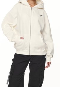 Donna che indossa una felpa con cappuccio zip-up di colore bianco sporco con un piccolo logo nero e pantaloni cargo neri con tasche e un'etichetta bianca.