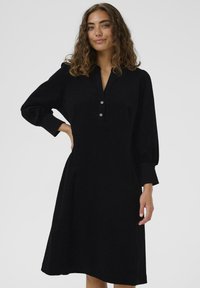 Black dress made of textured fabric, featuring a collared neckline, button detail, and long puffed sleeves, flaring skirts to knee length.