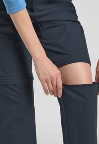 Knielange donkergrijze broek met een ritssluitingontwerp, waardoor aanpassing mogelijk is; gladde textuur; zichtbare hand die de stof vasthoudt.