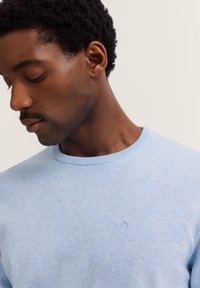 Nahaufnahme eines Mannes, der einen hellblauen Rundhalsausschnitt-Pullover mit dezentem gesticktem Logo auf der Brust trägt und gegen einen schlichten Hintergrund nach unten schaut.