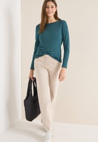 Camicia a maniche lunghe a righe color teal, pantaloni larghi color crema, tiene una borsa a sacco nera, sneakers bianche con dettagli rossi. Texture liscia.