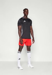 Črna športna majica s sivimi poudarki, oranžne kratke hlače, bele nogavice do kolen s črnimi črtami in beli športni čevlji.