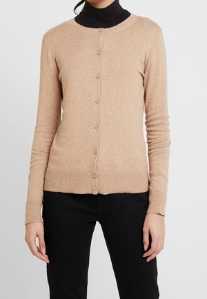 Femme portant un cardigan beige boutonné sur un col roulé noir et un pantalon noir, debout devant un fond clair uni.
