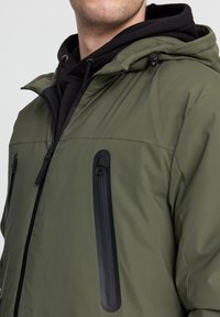 Giacca impermeabile verde con una tasca zip nera, texture morbida e cappuccio. Indossata sopra un felpa nera, evidenziando le zip e i dettagli.