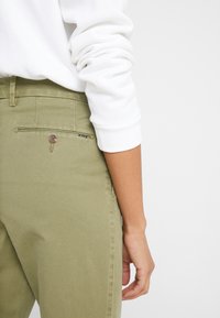 Green cotton pants with a smooth texture, featuring a back pocket with a zip and button detail, paired with a white long-sleeve top.