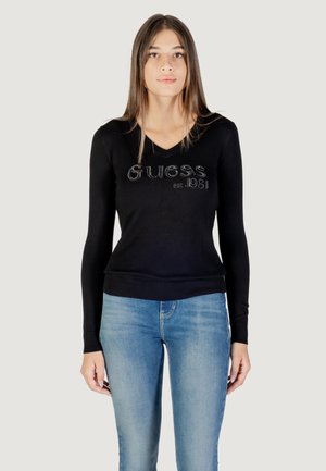 Maglietta nera a maniche lunghe con scollo a V, con la scritta "Guess est. 1981" in strass sulla parte frontale. Abbinata a jeans blu chiaro.