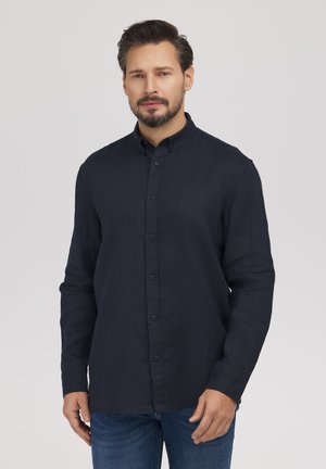 Mężczyzna o ciemnych włosach i brodzie, ubrany w zapiętą na guziki granatową koszulę i niebieskie dżinsy, stojący na prostym, jasnoszarym tle.