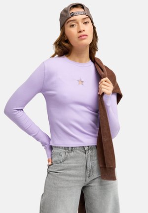 Young woman wearing a backward gray cap and lavender ribbed long-sleeve top with a star cutout, holding a brown sweater over her shoulder.