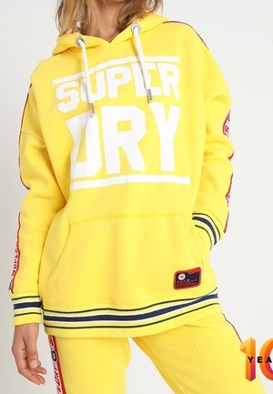 Person iført en lys gul hættetrøje og bukser med hvide snore og fed hvid tekst "SUPER DRY" på fronten.