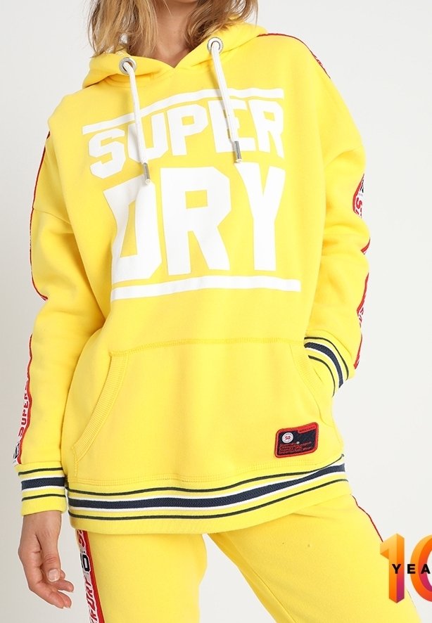 Personne portant un sweat à capuche jaune vif et un pantalon avec des cordons blancs et un texte blanc audacieux "SUPER DRY" à l'avant.