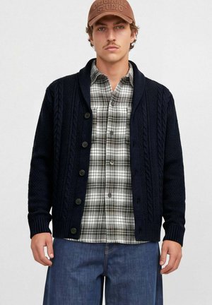 Un mannequin masculin porte une casquette marron, un cardigan bleu marine en maille torsadée, une chemise à carreaux noirs et blancs et un jean bleu, sur un fond clair uni.