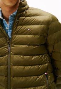 Oliven grønt quiltet jakke, med horisontal quiltedesign, høj krave og et lille logo patch. Lynlåslukning foran med kontrasterende detaljer.